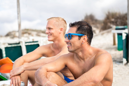 Two handsome young men chatting on a beach in their swimsuits sitting side by side on the sand with their backs to the ocean enjoying a relaxing summer day at the beachの写真素材
