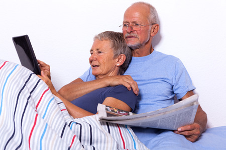 Senior Couple Relaxing in Bed with Tablet Computer and Newspaperの写真素材
