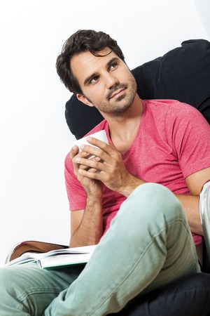 Young Man in Casual Clothing Sitting on Black Chair While Reading a Book and Holding a Glass of...