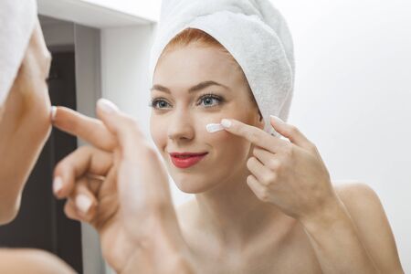 Close up Fresh Woman After Shower Applying White Cream on her Face in Front a Mirror With Towel on her Head.の写真素材
