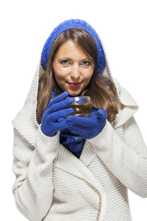 Fashionable young woman in a blue knitted winter ensemble and cowl neck jersey sipping a cup of hot tea with a smile in an effort to keep warm, isolated on whiteの写真素材