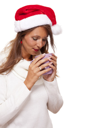 Cold attractive young woman with a cute smile in a festive red Santa hat sipping a hot mug of coffee that she is cradling in her hands to warm up in the winter weather, on whiteの写真素材