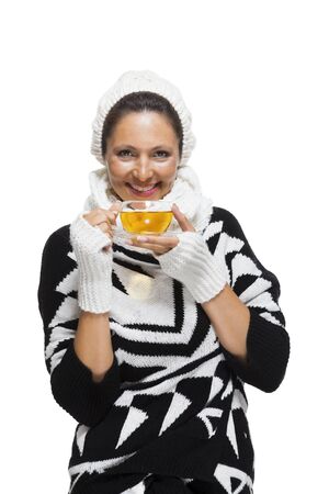 Elegant woman in a stylish black and white winter outfit with matching white mitts, scarf and knitted hat drinking a cup of hot tea, isolated on whiteの写真素材