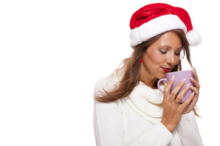 Cold attractive young woman with a cute smile in a festive red Santa hat sipping a hot mug of coffee that she is cradling in her hands to warm up in the winter weather, on whiteの写真素材