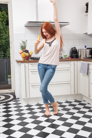 Happy Pretty Young Woman Holding a Banana Fruit, Dancing Alone at the Kitchen Inside the Home.の写真素材