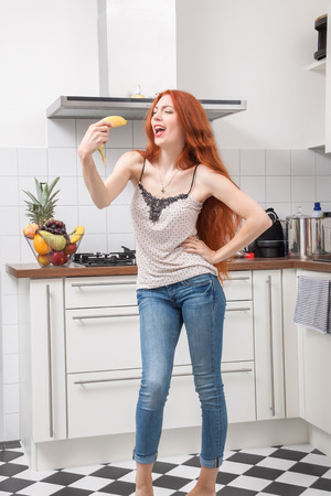 Happy Pretty Young Woman Holding a Banana Fruit, Dancing Alone at the Kitchen Inside the Home.の写真素材
