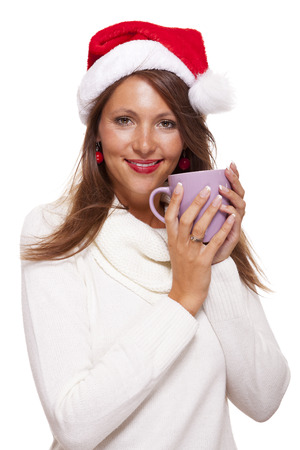 Cold attractive young woman with a cute smile in a festive red Santa hat sipping a hot mug of coffee that she is cradling in her hands to warm up in the winter weather, on whiteの写真素材