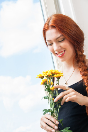 Close up Thoughtful Pretty Young Woman Holding Yellow Flowers, Looking Outside While Leaning on Glass Window.の写真素材