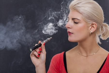 Stylish blond woman smoking an e-cigarette exhaling a cloud of smoke with her eyes closed in enjoyment, profile view on a dark backgroundの写真素材