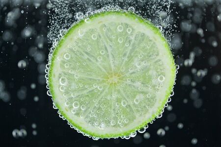 Close-up of single green lime slice falling into carbonated water with bubbles on black background. Refresher drink conceptの写真素材