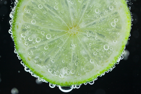 Close-up of single green lime slice falling into carbonated water with bubbles on black background. Refresher drink conceptの写真素材
