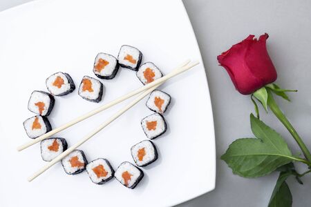 Sushi rolls laid out in the shape of a heart on a white plate. Red rose on a gray background. Creative greetings. The concept of Japanese cuisine for Valentine's Day, greeting card, banner. Copy spaceの写真素材