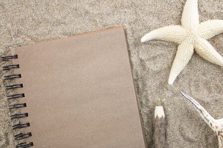 Notepad with copy space for text on the sea sand next to a starfish and shell, and eco wooden pen,in the sun. The concept of tourism, holidays and travel, vacation by the sea ocean. Flat lay.の写真素材