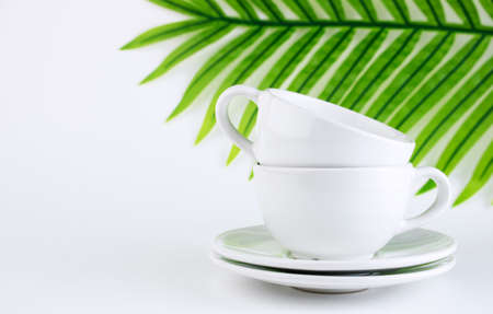 White mug and saucer on a white background, near palm leaves. A set of porcelain or ceramic dishes. Copy spaceの写真素材