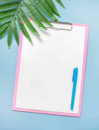 Minimalist concept of modern summer stationery layout. White empty blank, and pen, on a pink clipboard, on a blue background, near a palm leaf. Pastel shades. Top View. Flat Lay. Copy space.の写真素材
