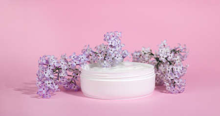 White jar of moisturizing face cream with pink cap isolated on pink background with lilac flowers. eco cosmetology or skin care concept.の写真素材