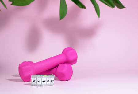 Measuring tape and pink dumbbells isolated on pink background with hard shadows and leaves. The concept of fitness, home workouts, activity and sports, healthy lifestyle. Women Health. Selective focusの写真素材