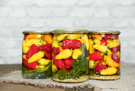 Mini chili peppers of red, yellow and orange colors, canned in glass jars with herbs, on a wooden table. Homemade pickled peppers in Asian style. Preparations for the winter. Copy spaceの写真素材