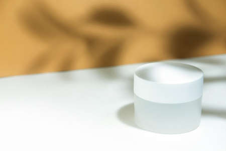 Empty white cosmetic jar container on a white beige background in the rays of the sun, with hard shadows from the razheniya. Cosmetology or skin care concept. Autumn cosmetics. Copy spaceの写真素材