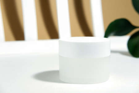 Empty white cosmetic jar on a white beige background in the rays of the sun, with harsh shadows from the plant. Cosmetology or skin care concept. Autumn cosmetics. Copy spaceの写真素材