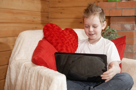 Valentine's Day children or kids. Cute little boy with a gadget, working or video chatting on a laptop, with a red heart pillow. Congratulations or postcard for Valentine's Day. Congratulations.の写真素材