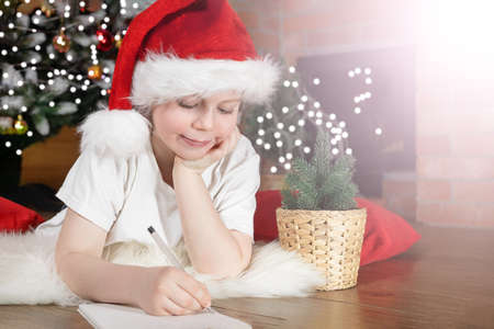 Happy Christmas kids. Boy child in Santa Claus hat writes a letter to Santa under the Christmas tree with Christmas lights, near the fireplace. Family Christmas.の写真素材