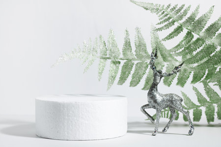 Creative christmas layout cylindrical podium and white green snow-covered leaves with crystals of snowflakes on a white background, near the deer, in the rays of the sun with shadows. Minimal empty cosmetic product presentation scene. Geometric podium. Christmas and New Year conceptの写真素材