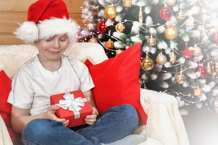 Merry Christmas kids. Emotions of a child. Joyful surprised kid boy in Santa's hat opened a Christmas box with a gift from Santa Claus near the Christmas tree with Christmas tree lights, near the fireplace. Family Christmas.の写真素材