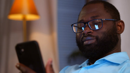 Smiling african american man holding smartphone using social media apps sitting on couch at homeの写真素材