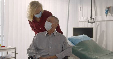 Senior wife in protective mask pushing discharged male patient on wheelchair in hospitalの写真素材