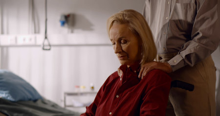 Portrait of pensive senior woman in wheelchair looking away with husband hands on her shouldersの写真素材