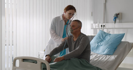 Woman doctor using stethoscope examining male aged patient with oxygen mask in hospital bedの写真素材