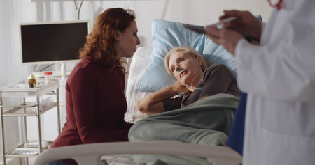 Senior woman patient lying in hospital bed with adult daughter sitting near and listening to doctorの写真素材