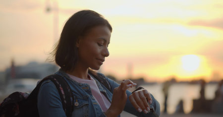 Bokeh shot of African-American woman traveler with backpack using smartphone outdoorsの写真素材