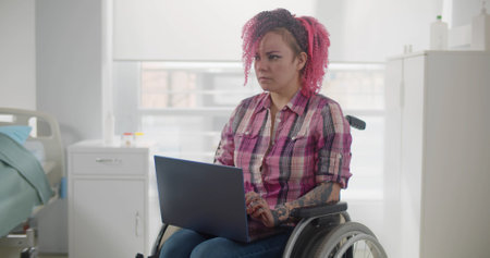 Disabled woman sitting in wheelchair and using laptop in hospital wardの写真素材
