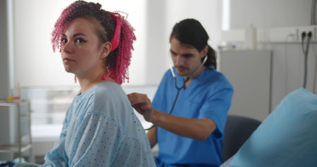 Male doctor using stethoscope to check lungs of female patient sitting in hospital bedの写真素材