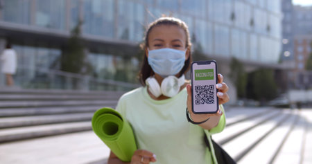 Sporty woman in safety mask looking at camera and showing smartphone with vaccination certificateの写真素材