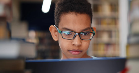 Close up of preteen african boy student smiling typing on laptop working at school library.の写真素材