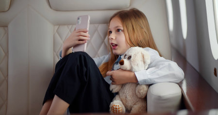 Adorable little girl with teddy bear talking on video chat on smartphone traveling by airplane.の写真素材