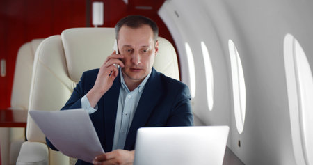 Mature businessman talking on phone and holding document sitting in private business planeの写真素材