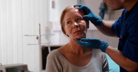 Male doctor examining wound on aged woman forehead in clinicの写真素材