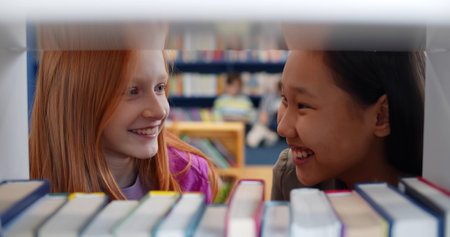 Teen girl students in library talking standing behind bookshelf