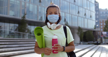 Young woman in protective mask holding yoga mat and bottle of water looking at camera outdoorsの写真素材
