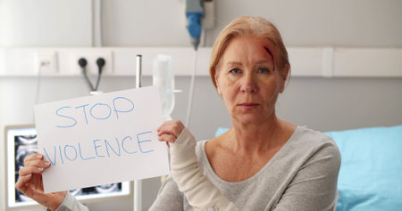 Senior woman with wounded forehead sitting on hospital bed and holding stop violence signの写真素材