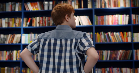 Back view of teen boy choosing book on bookshelf in libraryの写真素材