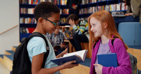 Multiethnic schoolchildren reading book and talking in libraryの写真素材