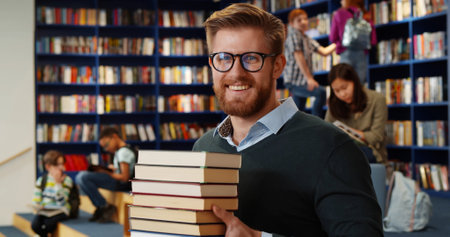 Bearded school teacher or librarian holding books in hands standing in library interior.の写真素材