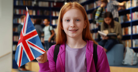 Pretty preteen girl holding great britain flag and smiling at camera standing in school libraryの写真素材