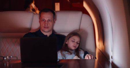 Businessman working on laptop and holding sleeping daughter flying on private jet at nightの写真素材