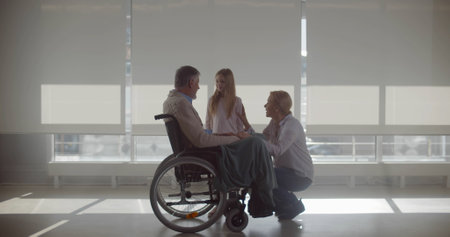 Senior man sitting in wheelchair and talking to wife and granddaughter near ag window at hospital corridorの写真素材
